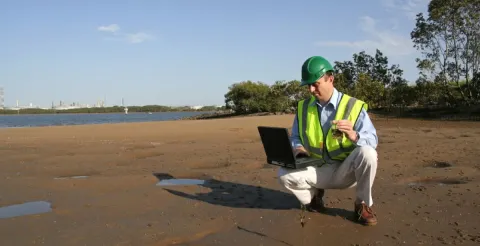 An environmental scientist stood on mudflats analysing data on his laptop