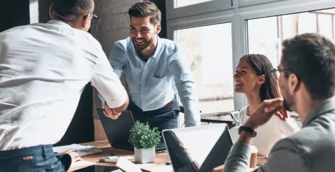 Group of professionals in a meeting room, one person stands and shakes hands with a colleague, while others smile and interact around laptops, suggesting leadership and teamwork