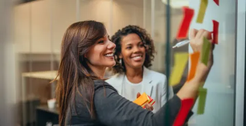 Two women smiling and collaborating while writing ideas on colorful sticky notes.