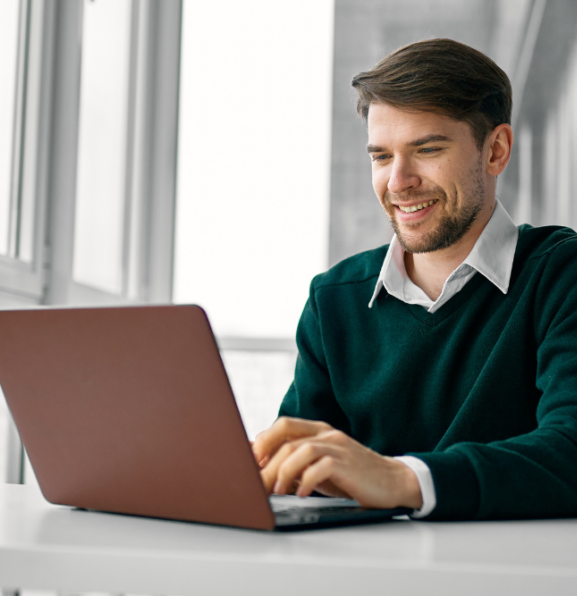 A man wearing a green sweater and white shirt smiles as he types on a maroon laptop at a white desk in front of a window.