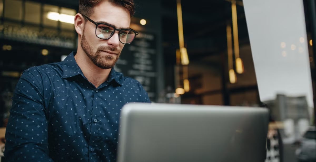 Young man with glasses works on a laptop in a modern café or workspace, representing starting a career in finance or entry-level job searches.