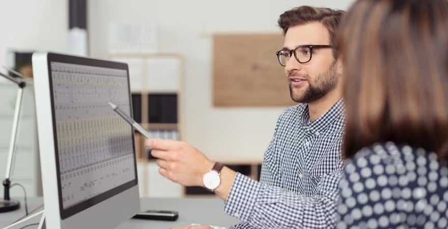 Two colleagues at a desk in an office review a large computer monitor displaying a spreadsheet, with one pointing at the screen with a pen while discussing data.