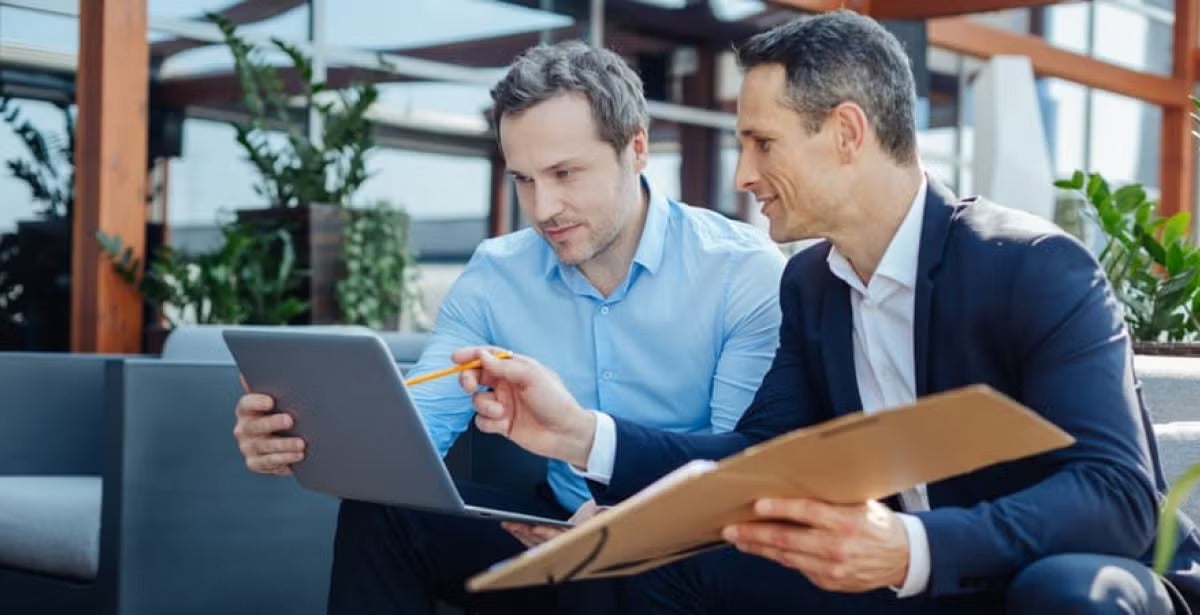 Two men in business attire sit together in a modern office lounge, discussing and reviewing information on a laptop and a folder.