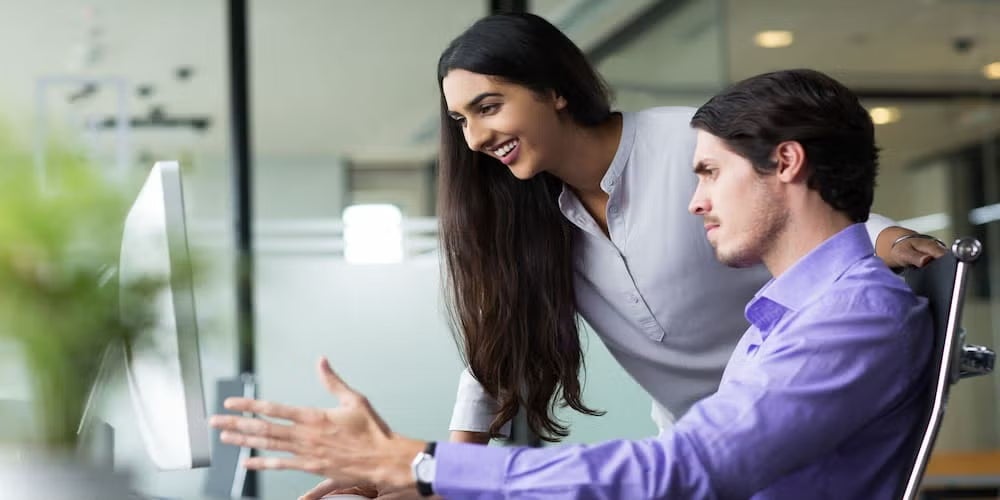 Man and woman collaborating at a computer in an office setting, underlining teamwork and knowledge sharing in cyber security roles.
