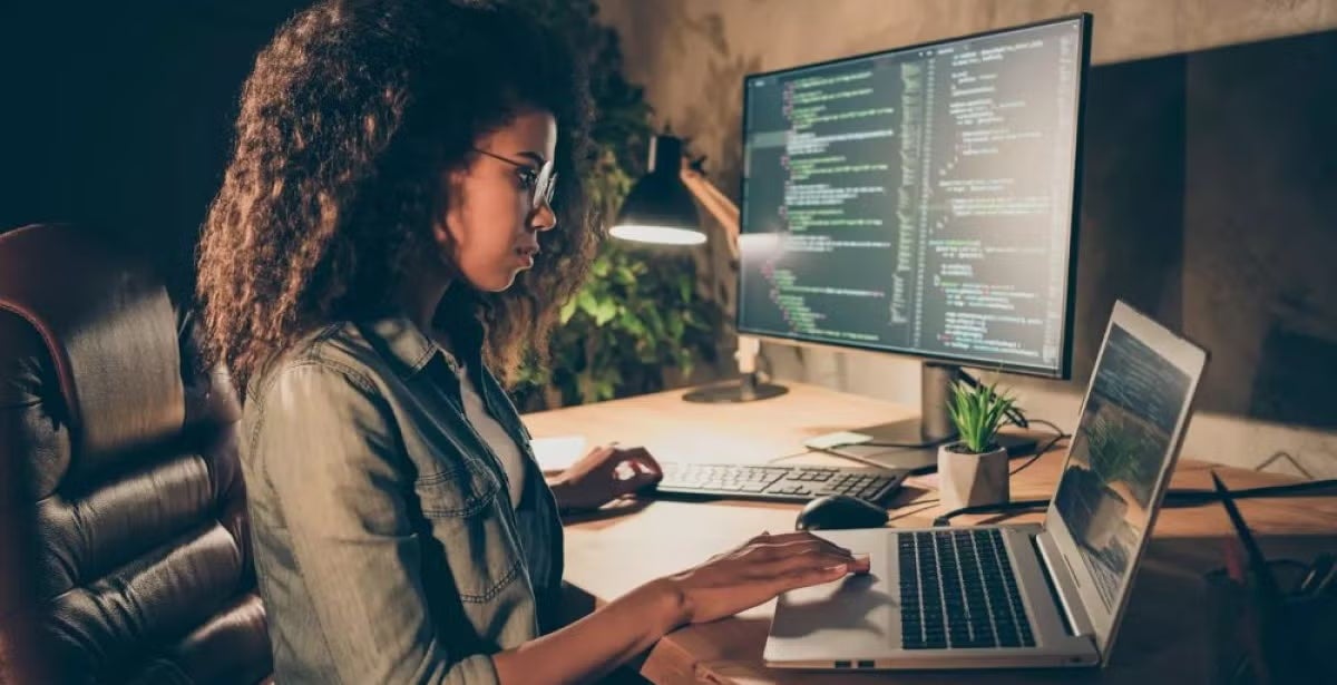 Woman with curly hair works at a desk with a laptop and large monitor displaying code, highlighting female representation in cybersecurity and tech.