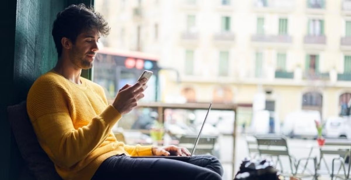 Young man in a yellow sweater sits by a window with a laptop, looking at his phone, suggesting remote work or digital job search in a city setting.