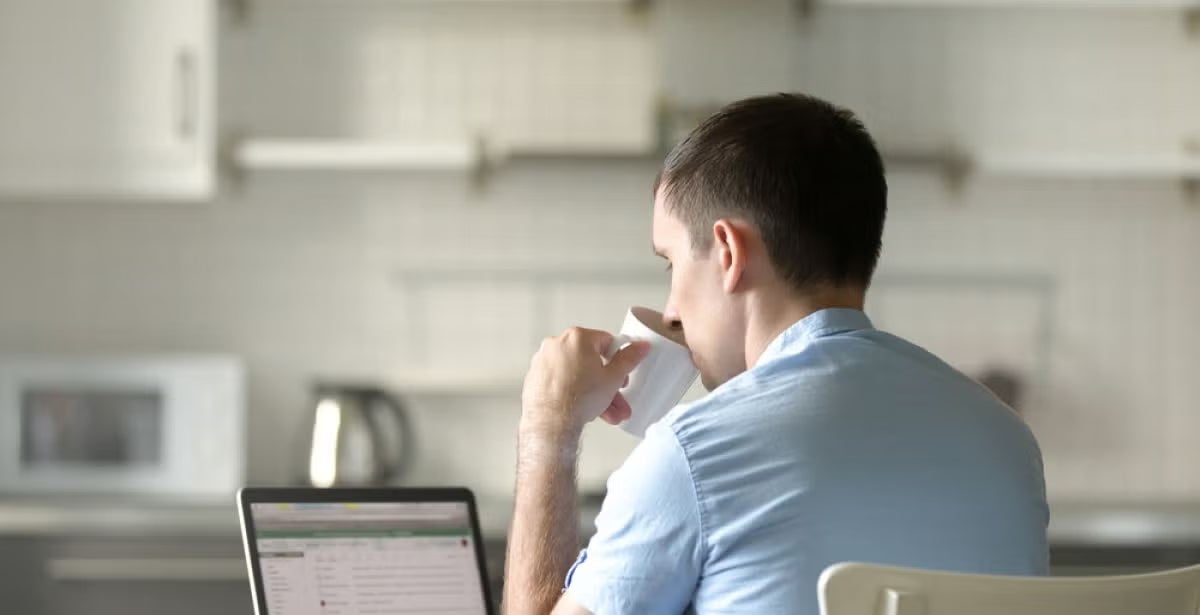 Man in a light blue shirt sits at a kitchen table with a laptop, drinking from a cup, suggesting remote learning or online study in a home environment.