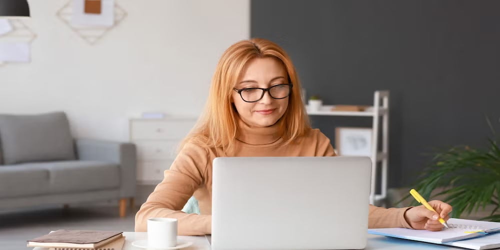 Woman in a tan sweater studies and takes notes with a laptop at a tidy desk, highlighting digital and applied learning.